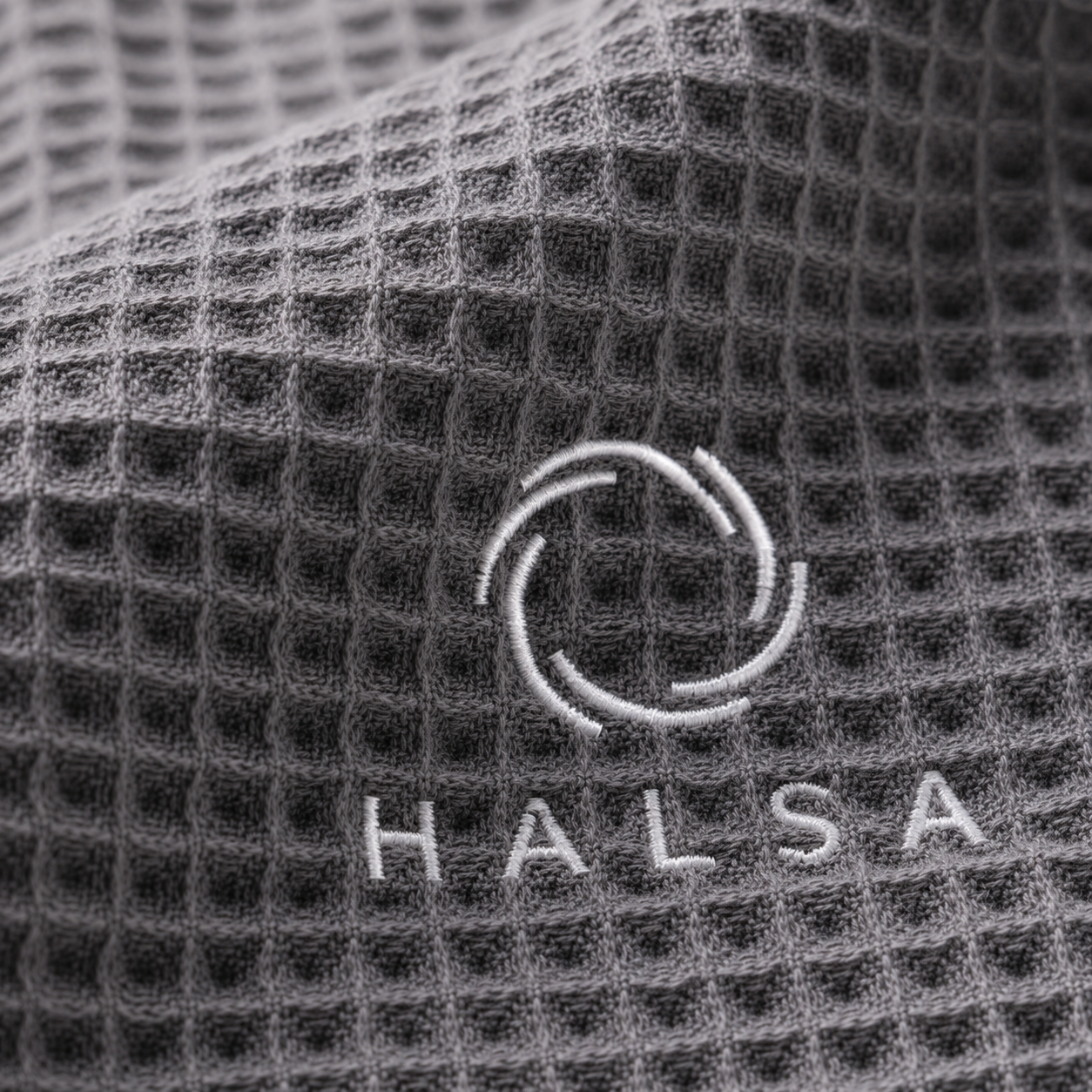 Macro close-up of the white embroidered HALSA logo stitched into the grey cotton waffle-weave sauna towel
