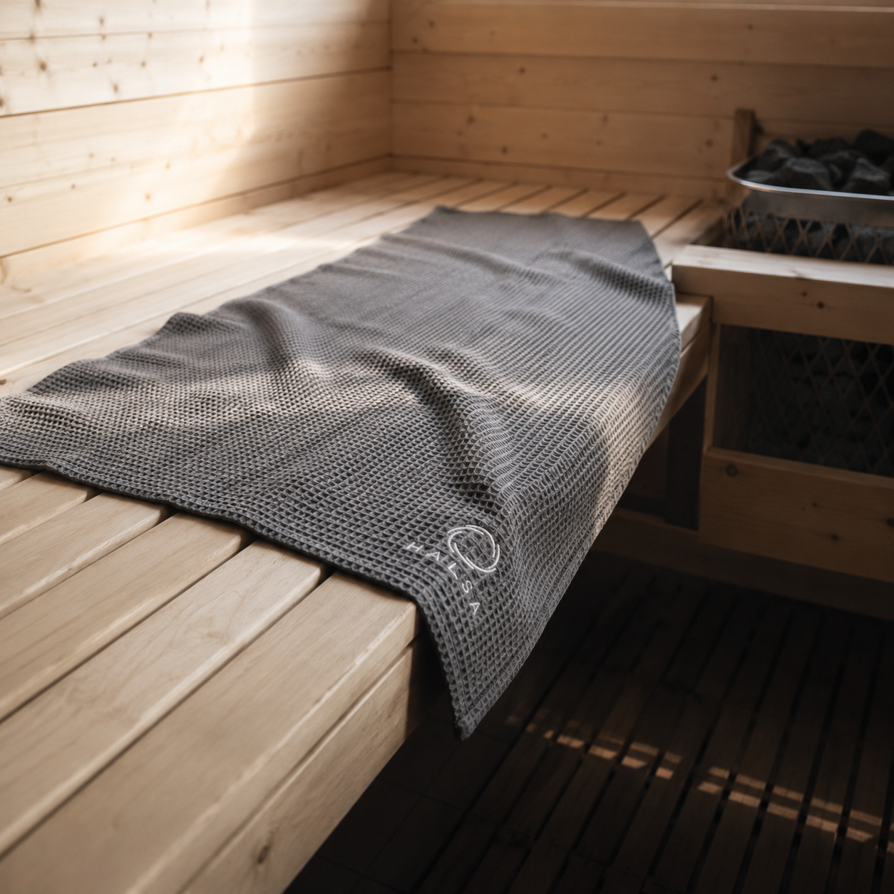 Halsa Waffle Sauna Towel draped across a Finnish sauna bench beside a sauna heater full of stones, with warm light through the wood