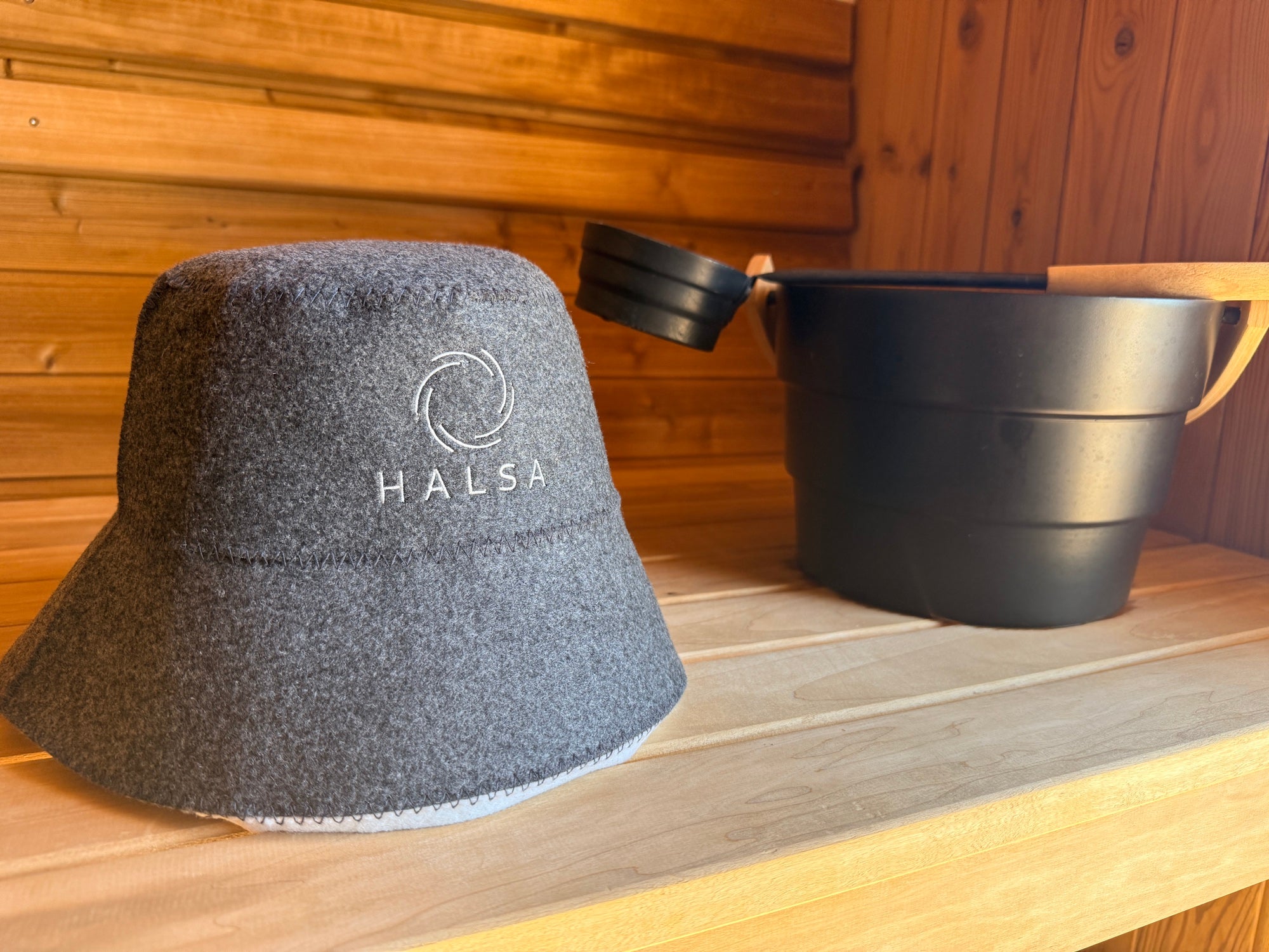 Halsa Bucket Wool Sauna Hat resting on cedar bench beside a black sauna bucket and ladle
